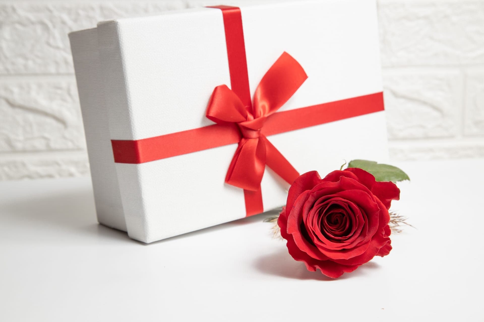 A white gift box with a red ribbon beside a single rose