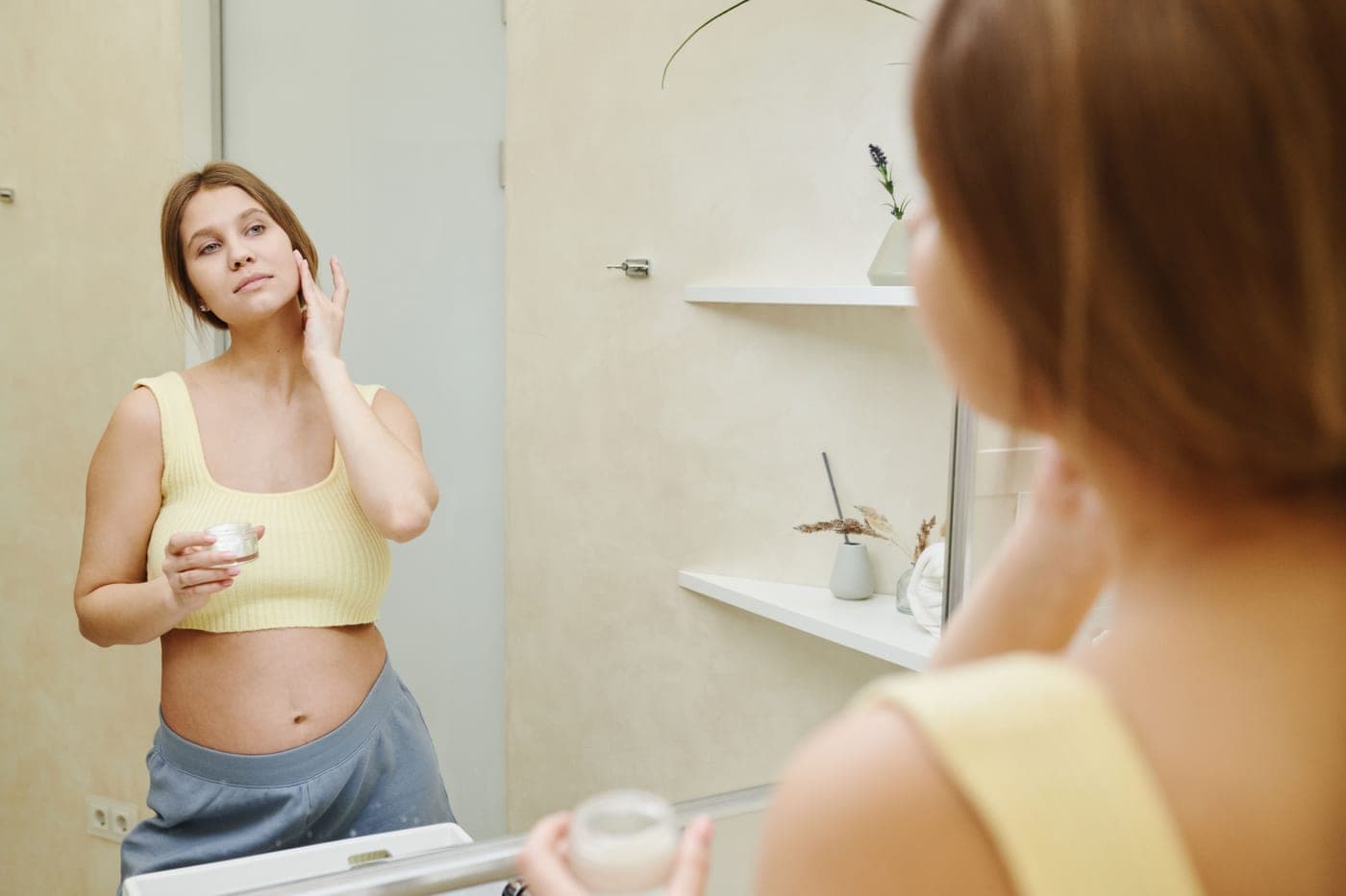A pregnant person applying moisturizer at a bathroom mirror