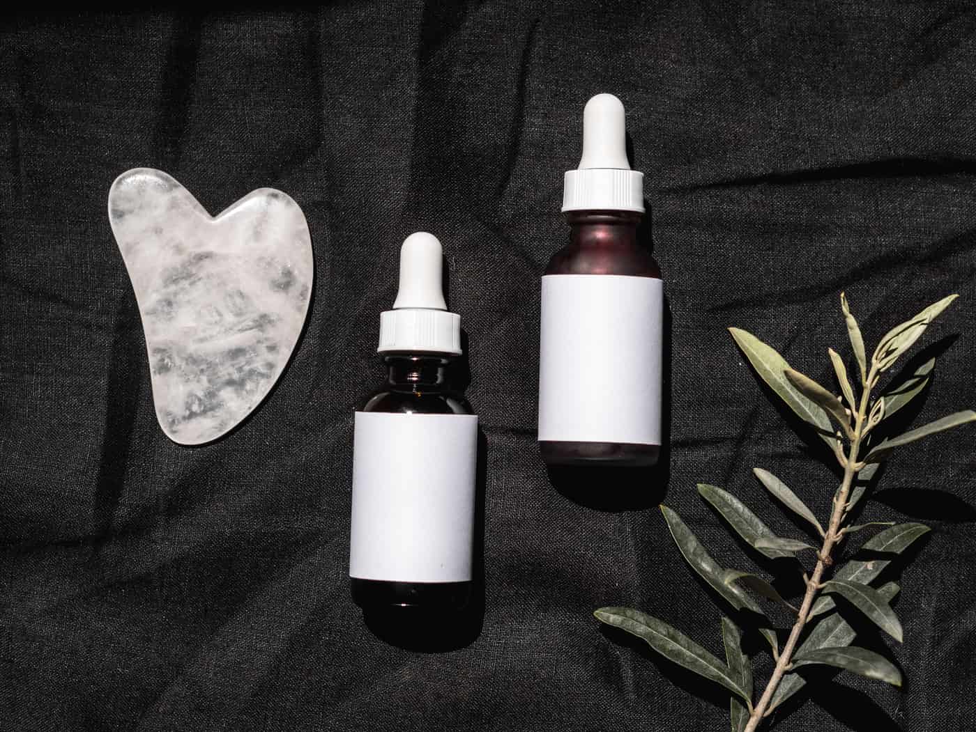 Two blank-label dropper bottles on dark linen beside a quartz gua sha and a sprig of leaves
